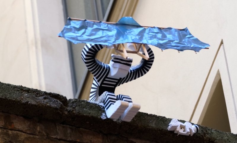 Small black-and-white striped figure holding a blue roof sculpture above a Berlin courtyard wall.